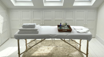 A white massage table with white towels and pitcher and glass of fresh water with lemon in a clean bright room bright with light flooding in through skylights.